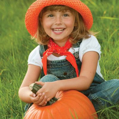 A little girl in overalls holding a pumpkin in a field.