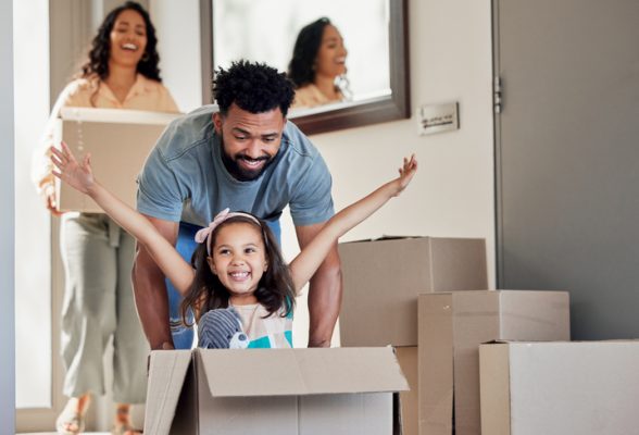 young father pushing excited daughter in moving box, mom smiles behind carrying another box as they move into new home.