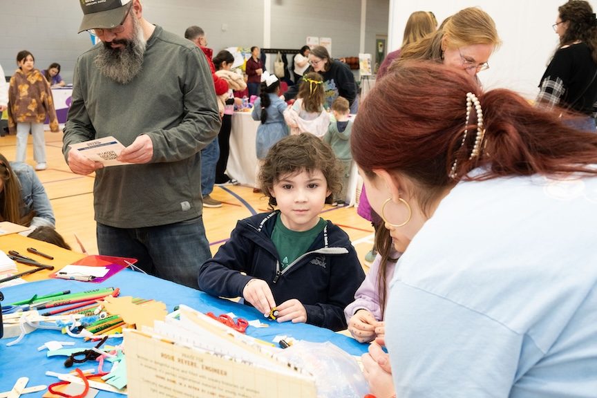 little kid smiles at camp booth as dad reads pamphlet behind him at CharlottesvilleFamily Camp Expo and KidFest